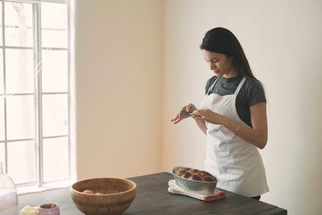 Woman in apron photographing baked Challah bread with mobile phone on table against wall at home Los Angeles, CA, United States ,model released, Symbolfoto CR_JAJEES200212-305476-01