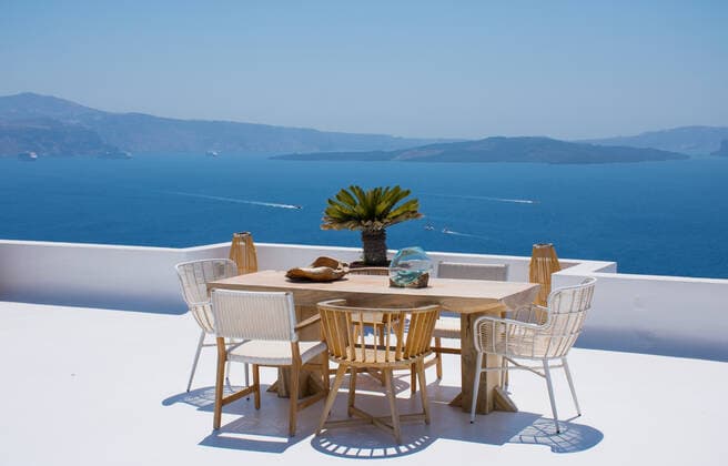 furniture composed by a table and some chairs on a white terrace of a house in Santorini Greece where you can enjoy meal while seeing a romantic seascape to the blue Aegean sea. Horizontal ph Oia, Greece CR_RUBO200219-310702-01