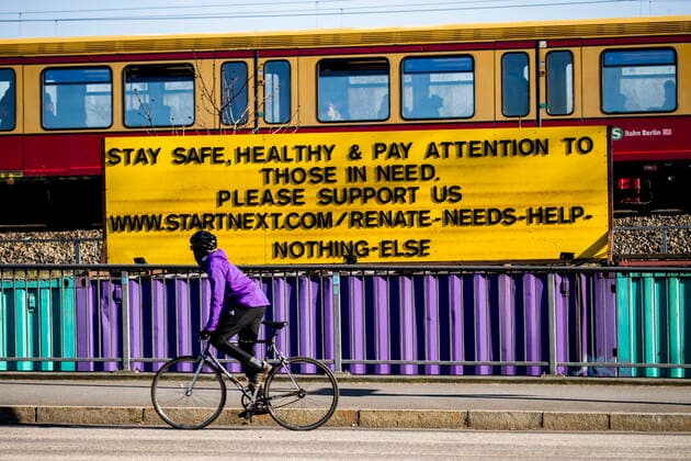 Ein Radfahrer faehrt vor einem Schild mit dem Schrift Stay safe, healthy and pay attention to those in need in Berlin am 23. Maerz 2020. Um die Verbreitung des Coronavirus (COVID-19) zu verlangsamen beschlossen Bund und Laender weitere Restriktionen. Nachdem Museen, Theatern, Sportveranstaltungen usw verboten wurden sind ab heute Versammlungen mit mehr als zwei Personen in der Oeffentlichkeit grundsaetzlich verboten. Restaurants koennen zum Beispiel nur nich Abholung anbieten. Oeffentliches Leber weiter in Berlin wegen Coronavirus eingeschraenkt
