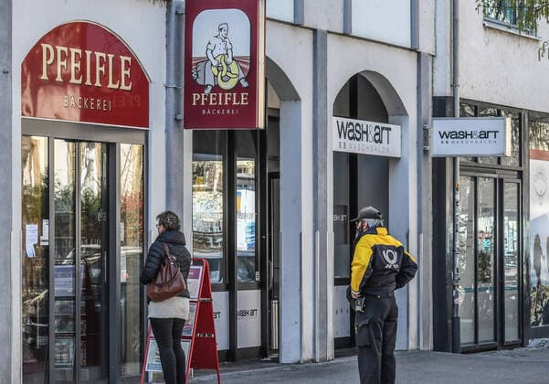 Ein Postbote steht in gebuehrendem Sicherheitsabstand an vor einer einer geoeffneten Baeckerei in einer sonst fast menschenleeren Stadt. Das oeffentliche Leben ist weitgehend heruntergefahren, Geschaefte und Läden geschlossen, Post, Muellabfuhr und andere systemrelevante Berufsgruppen sind weiterhin unterwegs. in Freiburg. Foto:Winfried Rothermel