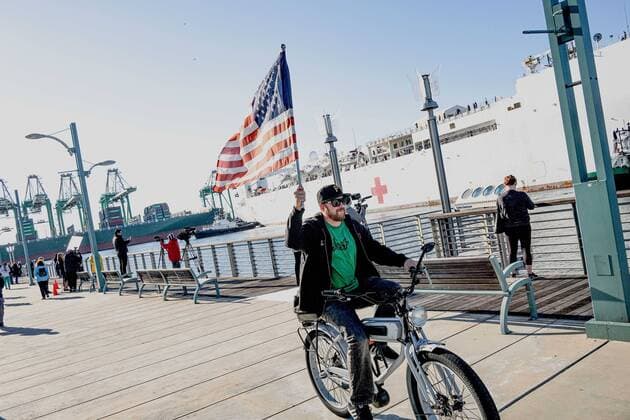 March 27, 2020, Los Angeles, California, USA: A man waves the US flag while riding his bike past Navy Hospital ship Mercy. The United States Naval hospital ship Mercy arrives in the port of Los Angeles as this will be the overflow hospital ship that will house patients not exposed to the ongoing Coronavirus pandemic. Los Angeles USA - ZUMAr145 20200327_new_r145_999 Copyright: xJasonxRyanx