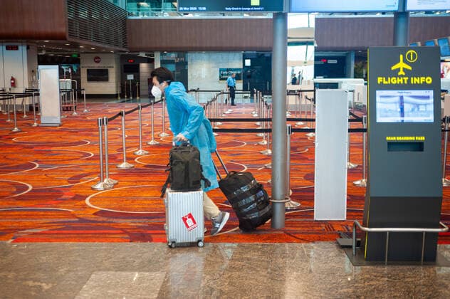 28.03.2020, Singapur, Republik Singapur, Asien - Ein Passagier kommt mit seinem Gepaeck an einem Abflugschalter im Terminal 1 am Flughafen Changi an und traegt eine Atemschutzmaske und einen Regenmantel, um sich vor einer Ansteckung mit dem pandemischen Coronavirus zu schuetzen. Der Flug mit China Southern Airlines nach Guangzhou (Kanton) ist einer von wenigen, die nach wie vor stattfinden nachdem es bereits zu massiven Flugausfaellen auf Grund der Coronakrise gekommen ist.