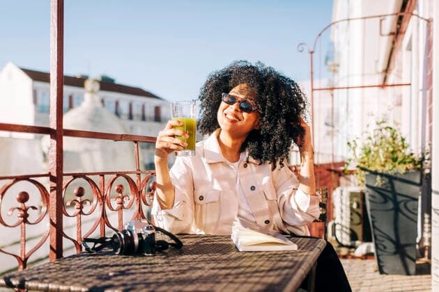 Smiling young woman with curly hair sitting at table on balcony with smoothie and camera model released Symbolfoto property released  DCRF00104
