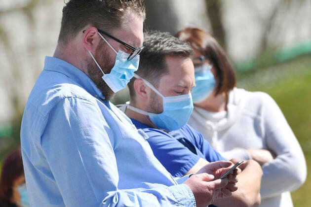 Personen mit Mundschutzmasken,Mundschutz, Masken,Schutzmasken.Nasenschutz,Mund-nasenschutz. Ein Mann schaut auf sein Smartphone,Tracking App,Tracing App.Handyapp. Zu viele Corona-Infektionen: Muenchner Helios Klinik gibt Alltagsbetrieb auf. Wegen einer Vielzahl von Corona-Infektionen unter Patienten und Mitarbeitern gibt die Muenchner Klinik den normalen Krankenhausbetrieb voruebergehend auf. Auch die Notaufnahme ist betroffen. So sollen Infektionsketten unterbrochen werden, am 02.04.2020