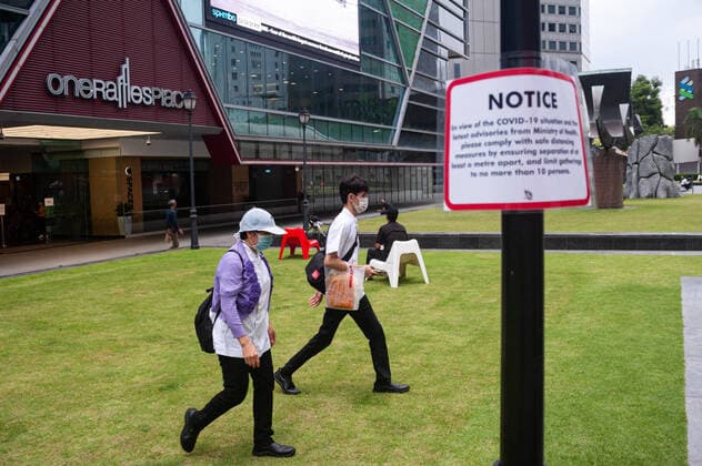 02.04.2020, Singapur, Republik Singapur, Asien - Menschen im Geschaeftsviertel am Raffles Place gehen an einem Hinweisschild zu Covid-19 vorbei und tragen einen Mundschutz, um eine Ansteckung mit dem pandemischen Coronavirus zu verhindern. Um die Ausbreitung des Coronavirus weiter einzuschraenken sind Massnahmen zur Einhaltung von Sicherheitsabstaenden (Safe Distancing) im oeffentlichen Leben eingefuehrt worden, was zur raeumlichen Distanzierung (Social Distancing) fuehrt und somit den unmittelbaren Kontakt mit Mitmenschen verhindern soll.