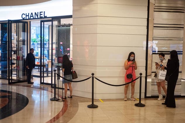 02.04.2020, Singapur, Republik Singapur, Asien - Frauen stehen vor einem Geschaeft der Luxusmarke Chanel im Takashimaya Einkaufszentrum an und tragen dabei eine Atemschutzmaske, um sich vor einer Ansteckung mit dem pandemischen Coronavirus zu schuetzen.