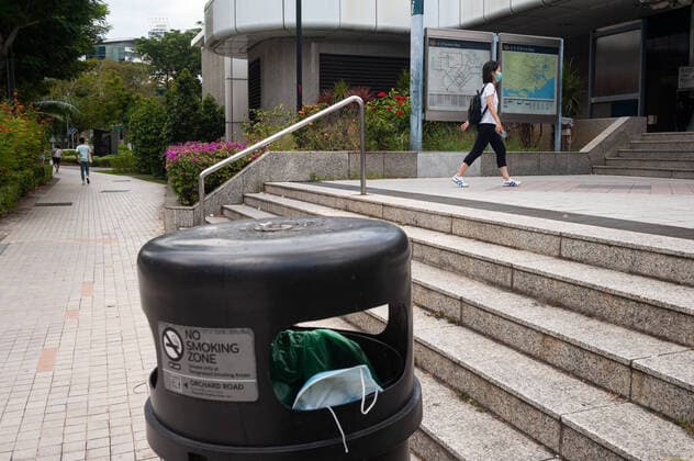 02.04.2020, Singapur, Republik Singapur, Asien - Ein weggeworfener Mundschutz haengt in einem Muelleimer vor einer U-Bahnstation in der Innenstadt.