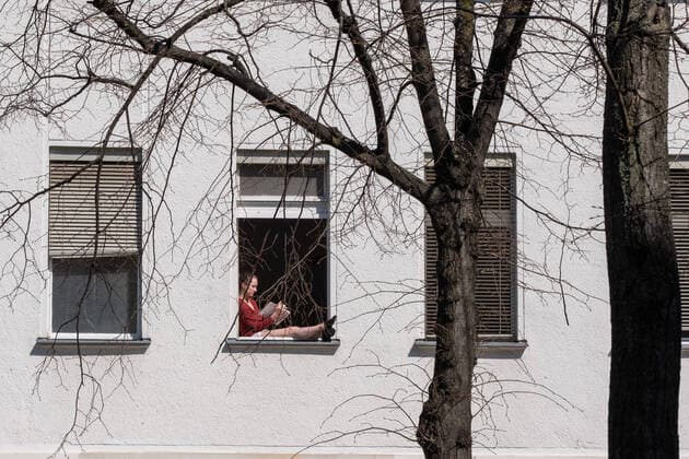 Ausgangsbeschränkung & Kontaktverbot - Frau sitzt in ihrem Fenster in Berlin-Prenzlauer Berg und liest bei warmen Frühlingswetter ein Buch Coronakrise - Ausgangsbeschrämkungen