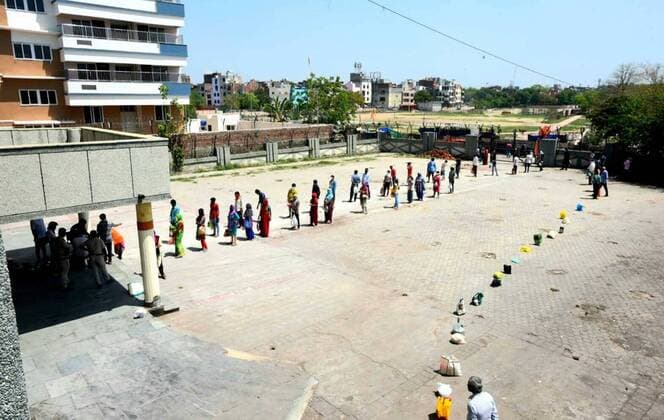 NEW DELHI, INDIA - APRIL 6: People queue while keping distance at a food distribution by the Delhi Government at a Basti Vikas Kendra, on day thirteen of the 21-day nationwide lockdown to check the spread of coronavirus at Gandhi Nagar on April 6, 2020 in New Delhi, India. A queue on the right is filled with placeholders while the people shelter under a shed, violating social distance. (Photo by Sonu Mehta Hindustan Times) Government And NGOs Provide Food To Homeless And Poor Amid Lockdown To Prevent Covid-19