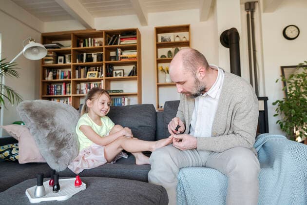 IMAGO Creative: Vaterschaft Father painting his daughter s toenails with red nail polish on the couch in living room model released Symbolfoto property released VYF00090