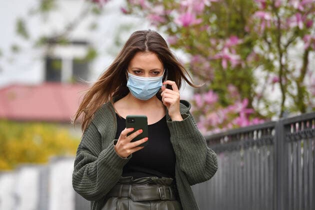 Themenbild Corona: Eine junge Frau mit Schutzmaske,Mundschutz, Community Maske blickt auf ihr Smartphone am 24.04.2020.