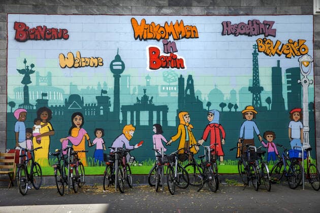 IMAGO Creative: Kollidierende Kulturen Fahrraeder stehen vor einem Wandgemaelde mit der Skyline von Berlin und der Aufschrift Willkommen in Berlin in verschiedenen Sprachen vor dem Landesamt fuer Einwanderung in Berlin, 07.07.2020. Berlin Deutschland *** Bicycles stand in front of a mural with the skyline of Berlin and the inscription Welcome to Berlin in different languages in front of the State Office for Immigration in Berlin, 07 07 2020 Berlin Germany Copyright: xJaninexSchmitz photothek.netx