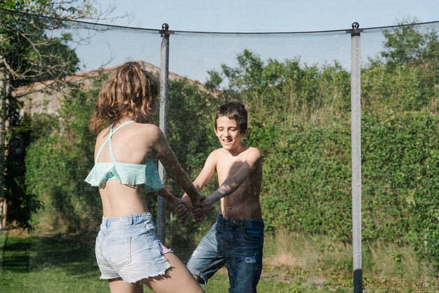 IMAGO Creative: Sommer, Sommertage im Garten Horizontal photo of a blonde boy and a brunette girl holding hands playing on a trampoline on a green playground. The teenager and the child wear short jeans and they look happy, smiling San Martín de la Virgen del Moncayo, Aragón, Spain CR_RUBO200708-448488-01 ,model released, Symbolfoto