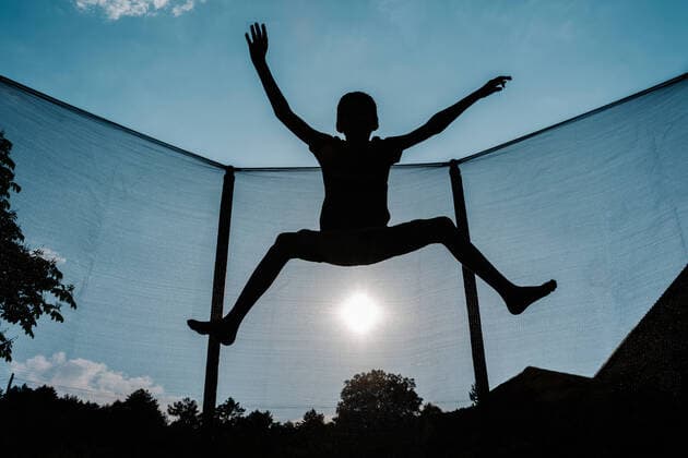 Horizontal front backlighting photo of a silhouette of a barefoot young boy jumping or flying on a trampoline with net and the sun at the background reflecting sunbeam rays on his shadow San Martín de la Virgen del Moncayo, Aragón, Spain CR_RUBO200708-448531-01 ,model released, Symbolfoto