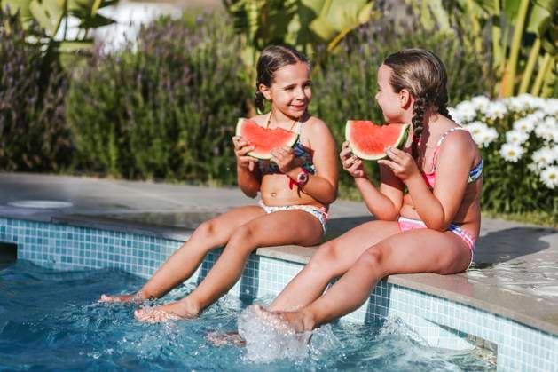 Happy twin girls sitting with watermelon slice at poolside on sunny day model released Symbolfoto property released LJF01574