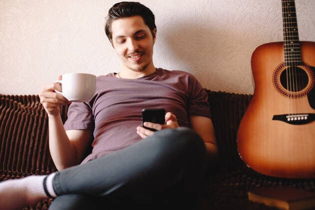 Happy young man using smart phone while drinking tea at home CR_DMBE200812-379179-01 ,model released, Symbolfoto