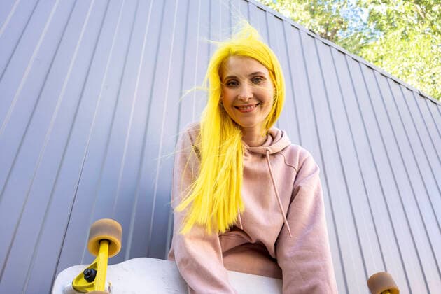 Smiling dyed yellow hair woman standing with skateboard against metal wall model released Symbolfoto VPIF03130