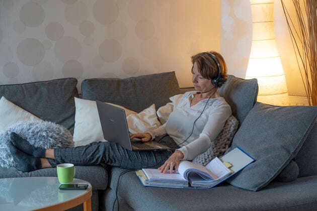 Frau, Mitte 50, arbeitet von Zuhause aus, mit Laptop und kommuniziert per Headset mit Kollegen, Homeoffice, auf dem Sofa, Homeoffice