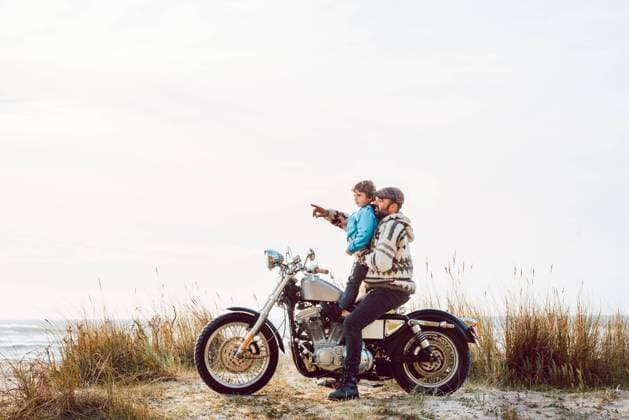 Side view of adult bearded man in sweater and little boy sitting together on motorcycle spending time on ocean shore, Model released Copyright: xAlbertoxMenendezx MR