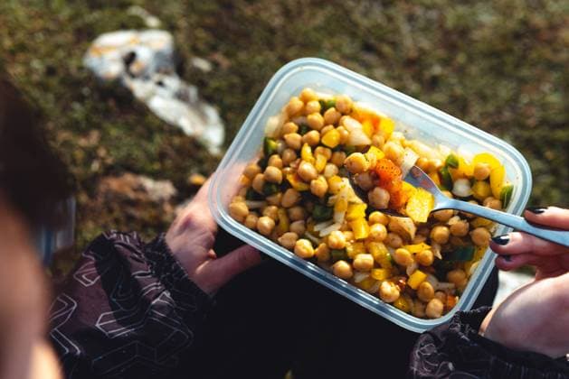 From above crop woman in warm jacket holding plastic container of chickpea with veggies having lunch on meadow while hiking in Spain, Model released Copyright: xAlbertoxMenendezx MR