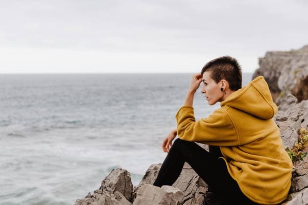 IMAGO Creative: Gesundheit, Stille Side view of traveler woman in yellow warm hoodie sitting alone on rocky shore looking at foamy waves on cloudy day, Model released Copyright: xAlbertoxMenendezx MR