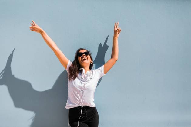 Carefree beautiful woman in casual outfit and headphones on neck standing with hands up beside blue wall of building on city street, Model released Copyright: xAlexandraxCoelhox MR