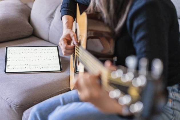 IMAGO Creative: Bildung, Musikerziehung Woman playing guitar sitting on her couch at home and learning with online lessons with a digital tablet with a digital tablet with a blank screen from above, Model released, Property released Copyright: xAlvaroxSanchezx MR