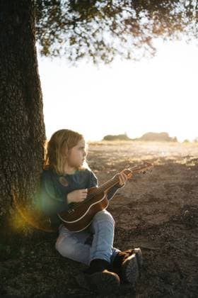 Adorable serious little girl in casual wear playing ukulele guitar while sitting near tree in sunny summer day in countryside Copyright: xAngelxSantamariax Model Released