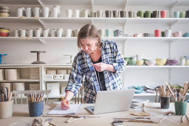 Mature female potter writing on paper while standing by laptop at table in workshop , model released, property released Copyright: xMaskotx , MA55813