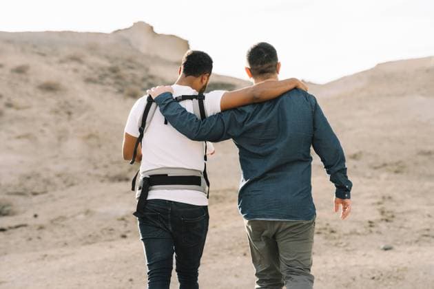 IMAGO Creative: Pride Month 2022, Engagement der Jugendlichen Back view of cropped black casual man holding on shoulder anonymous male boyfriend and holding little calm baby in grey carrier while strolling on nature at daytime, Model released Copyright: xGabrielxTrujillox MR