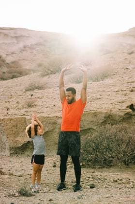 IMAGO Creative: NGO, Ausbildung f?r Kinder Side view of African American bearded man in activewear standing opposite curly toddler teaching stretching front of thigh on sandy landscape, Model released Copyright: xGabrielxTrujillox MR