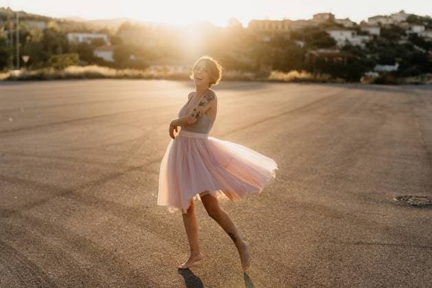 IMAGO Creative: Lokalhelden - Kunst und Kultur Full body delighted young female artist in elegant dance dress laughing and dancing in back lit in empty square with blurred town in background in sunny summer sunset day Copyright: xGustavoxPozox Model Released