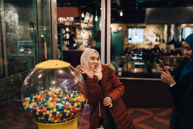 IMAGO Creative: Kollidierende Kulturen Young woman photographing cheerful female friend standing with bubble gum by vending machine , model released Copyright: xMaskotx , MA61998