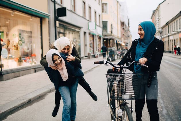 Young woman walking with bicycle looking at cheerful teenage girl giving piggyback to friend on street in city , model released Copyright: xMaskotx , MA62076