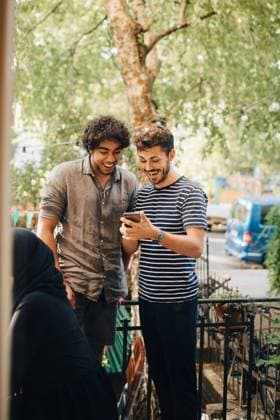 Cheerful young man showing smart phone to male friend while standing in balcony during party , model released, property released Copyright: xMaskotx , MA68046