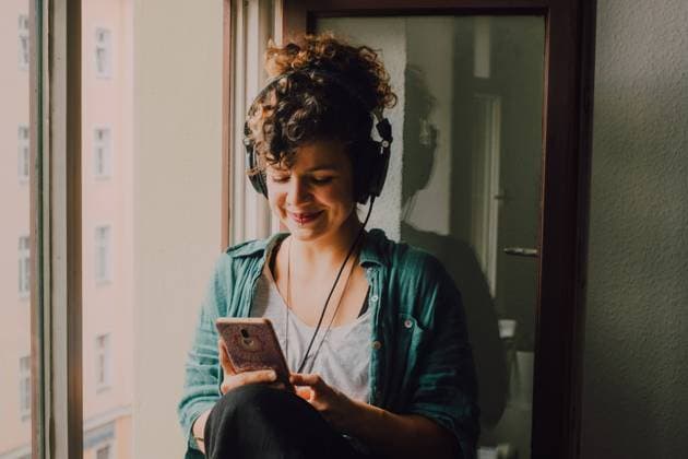 IMAGO Creative, Lifestyle, Tik Tok-Trend thatgirl Smiling curly woman in headphones listening to music while browsing smartphone and sitting on window sill in apartment, Model released Copyright: xJakexJakabx MR