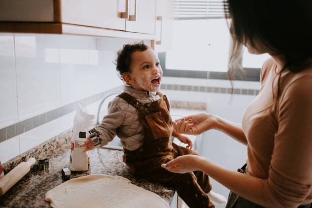 Side view of cheerful little boy in casual clothes with flour on cheeks helping mother cooking pastry in modern kitchen, Model released Copyright: xJakexJakabx MR
