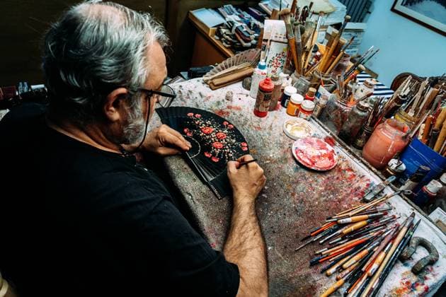 IMAGO Creative: 2021 Favoriten Painter using paintbrush to draw on hand fan while sitting at messy table and working in creative workshop Copyright: xJavierxBallesterx Model Released