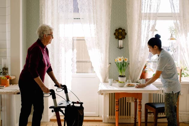 IMAGO Creative: Zivildienst und ehrenamtliches Engagement Retired senior woman walking with rollator while young female caregiver serving breakfast on table in nursing home , model released, property released Copyright: xMaskotx , MA70858