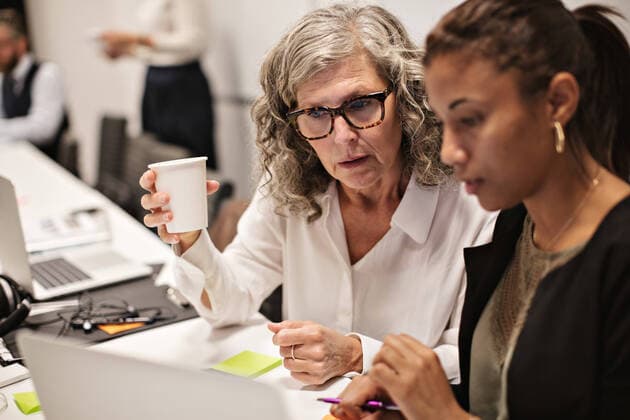 Senior businesswoman having coffee while discussing with colleague over laptop at office , model released, property released Copyright: xMaskotx , MA70901