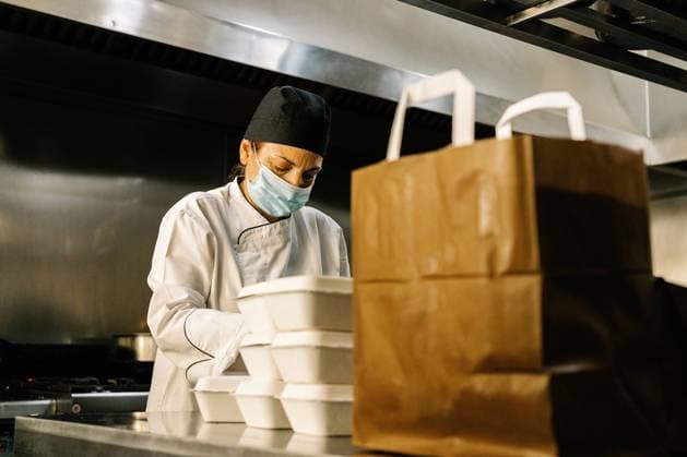 Concentrated female chef wearing face mask and uniform while working in modern restaurant kitchen and packing containers with food into paper bag for takeaway Copyright: xJavierxBallesterx Model Released