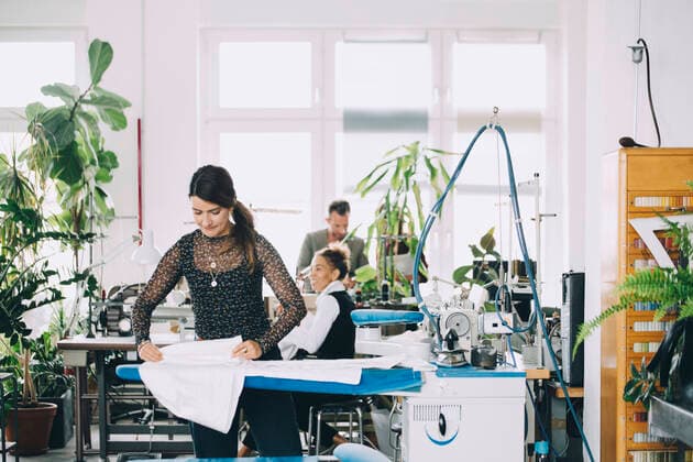IMAGO Creative: Kreative Renaissance -- der junge Entrepreneur Female entrepreneur ironing white fabric while colleagues working in background at creative office , model released, property released Copyright: xMaskotx , MA72905