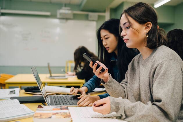 Female high school student using smart phone while sitting by classmate studying on laptop in classroom , model released, property released Copyright: xMaskotx , MA73310