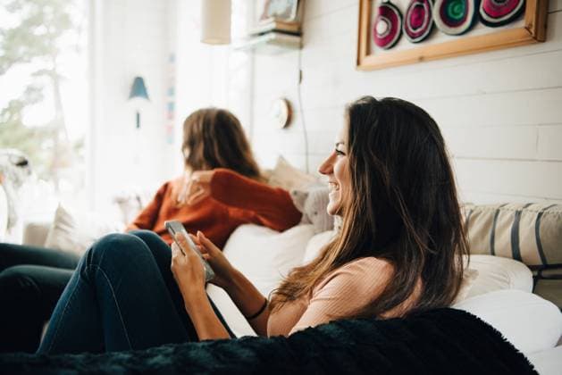 Smiling woman with smart phone looking away while sitting with friend on sofa at home , model released, property released Copyright: xMaskotx , MA75221