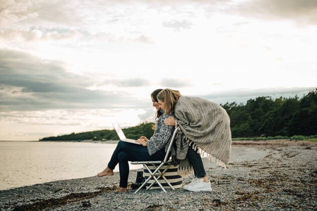 IMAGO Creative: Landleben - Alfresco, Draußen Friend greeting woman working on laptop while sitting at beach against sky , model released Copyright: xMaskotx , MA75240