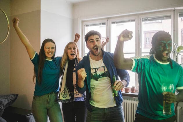 IMAGO Creative: Sport Start-Up IMAGO Creative: Frauen im Leistungssport Young cheerful friends watching soccer match while standing in living room , model released, property released Copyright: xMaskotx , MA77362