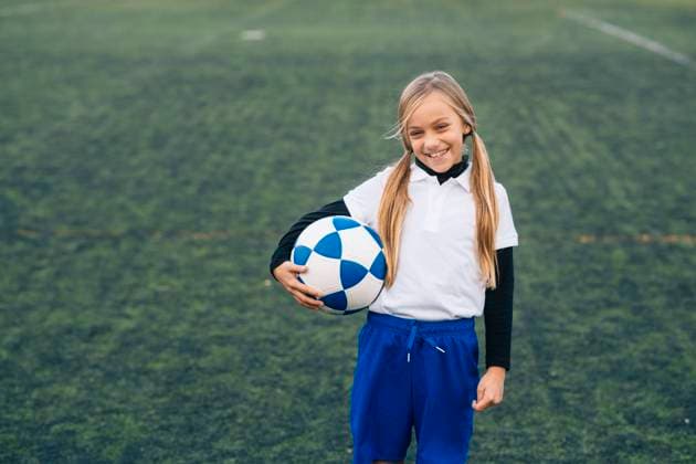 IMAGO Creative: Sport Start-Up Cheerful preteen girl in white and blue uniform with soccer ball smiling at camera while standing alone on green field in modern sports club, Model released Copyright: xJosexLuisxCarrascosax MR