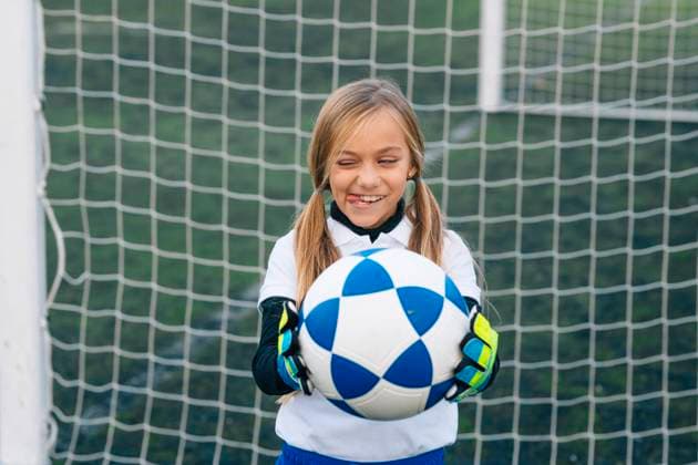 Playful preteen goalkeeper in uniform and gloves sticking out tongue while preparing to perform feint standing at goal post with ball in soccer arena in sports club, Model released Copyright: xJosexLuisxCarrascosax MR