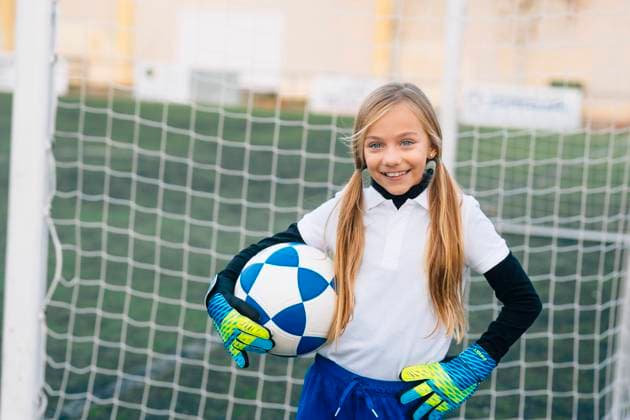 Cheerful preteen girl in white and blue uniform with soccer ball smiling at camera while standing alone on green field in modern sports club, Model released Copyright: xJosexLuisxCarrascosax MR