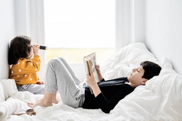 IMAGO Creative: Jahreszeit Winter, Fantasievoll drinnen spielen Side view of brother reading book and sister playing with retro spyglass relaxing on comfortable bed and enjoying weekend, Model released Copyright: xJuanxGarciaxRisquezx MR